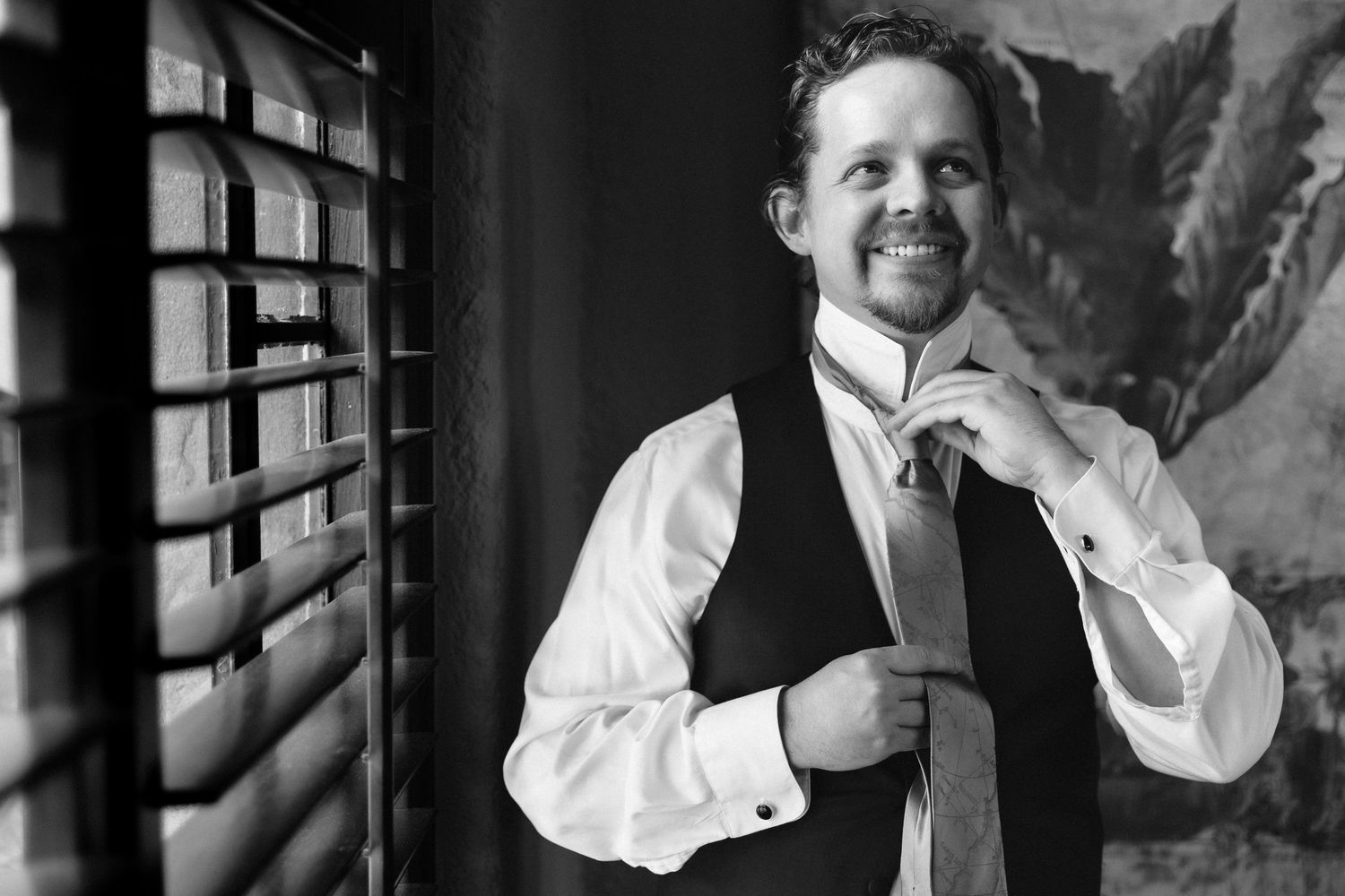 Dapper groom in navy vest adjusting his tie while smiling in a black and white wedding preparation photo.
