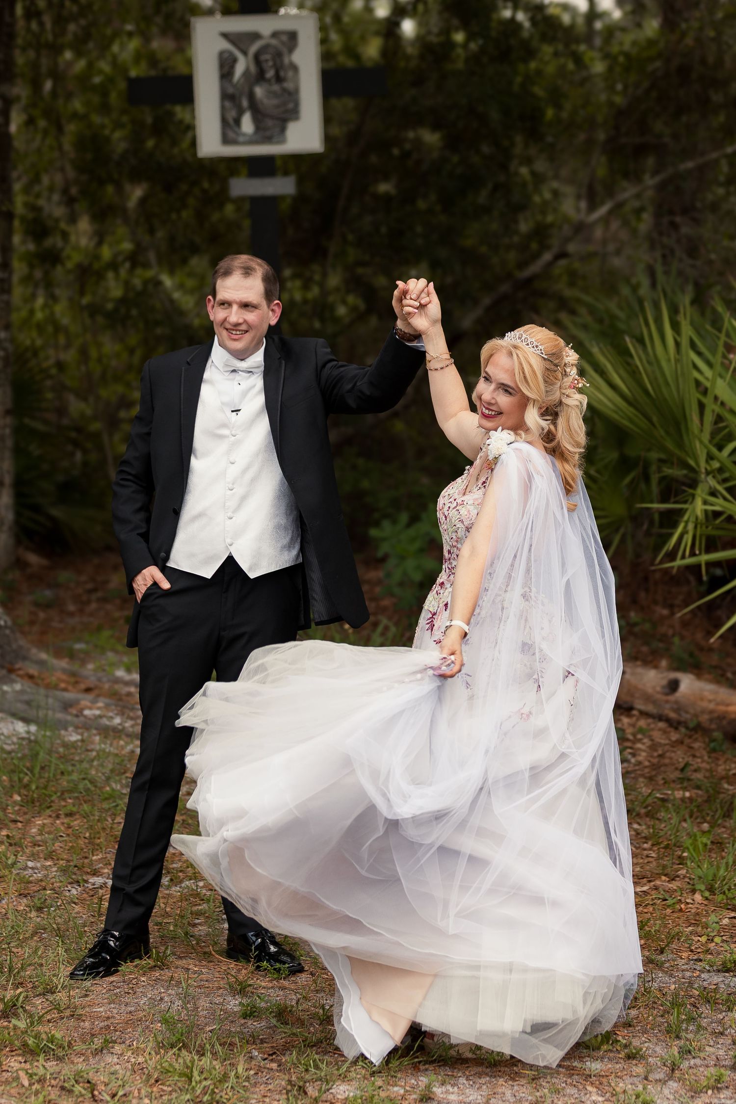 Groom twirling bride in floral wedding gown outdoors near religious cross surrounded by lush greenery.