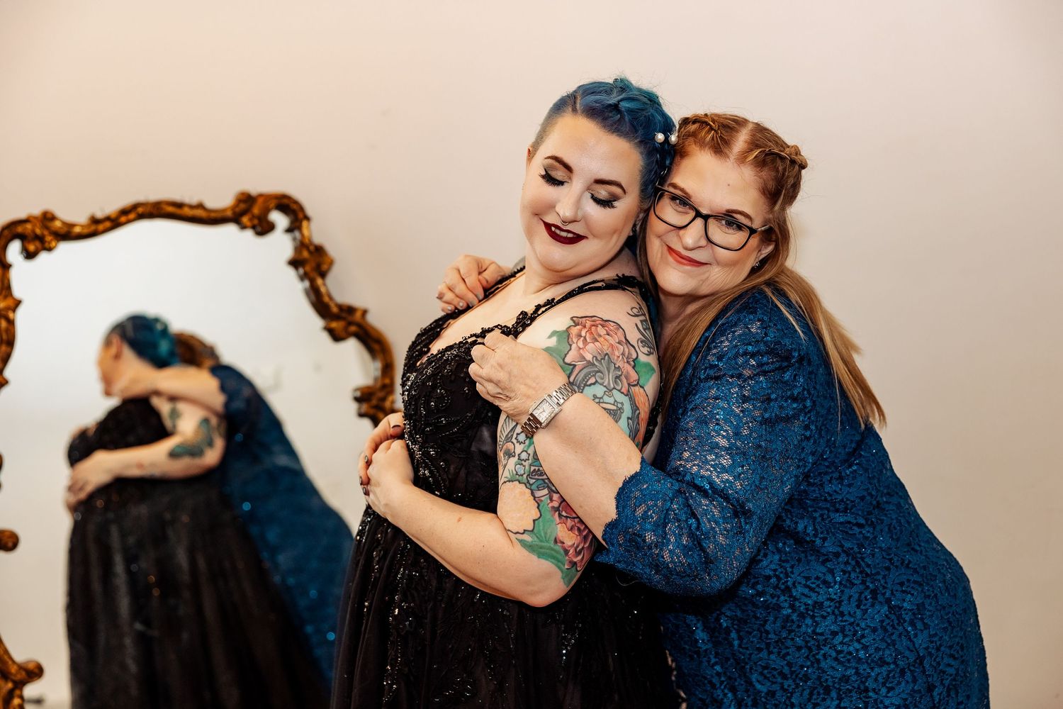 Tattooed bride in black gown embracing red-haired woman near ornate gold mirror.