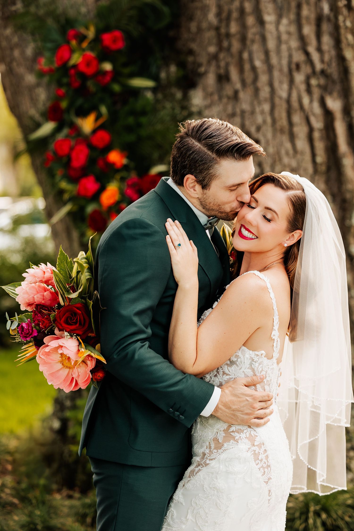 Groom kisses smiling bride on cheek outdoors near large tree with red floral arrangement backdrop.