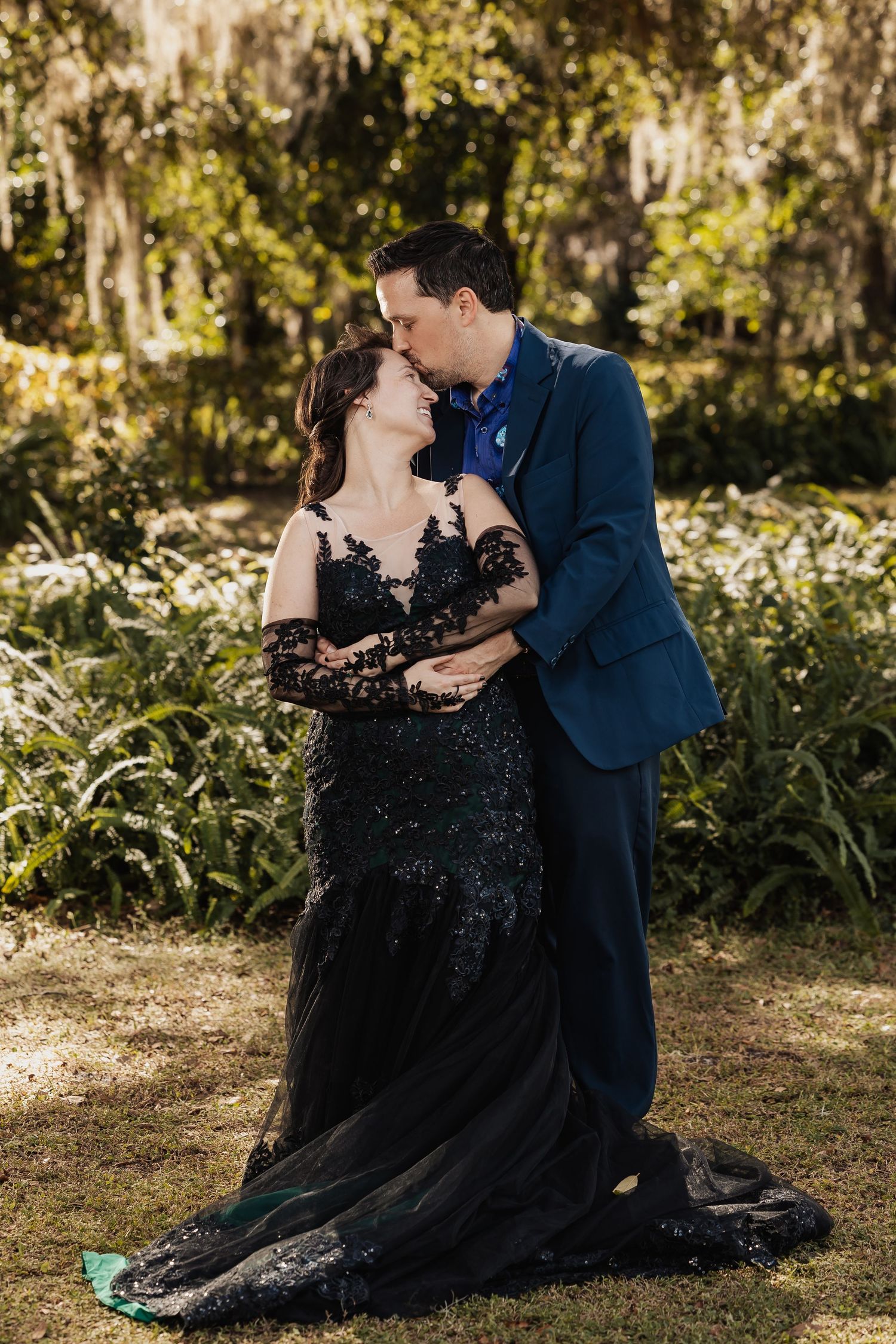 Bride in black lace gown and groom in blue suit sharing a romantic kiss in a sunlit garden setting.