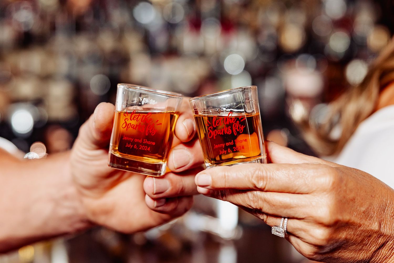 Close-up of bride and groom clinking whiskey glasses with wedding ring visible during reception toast.