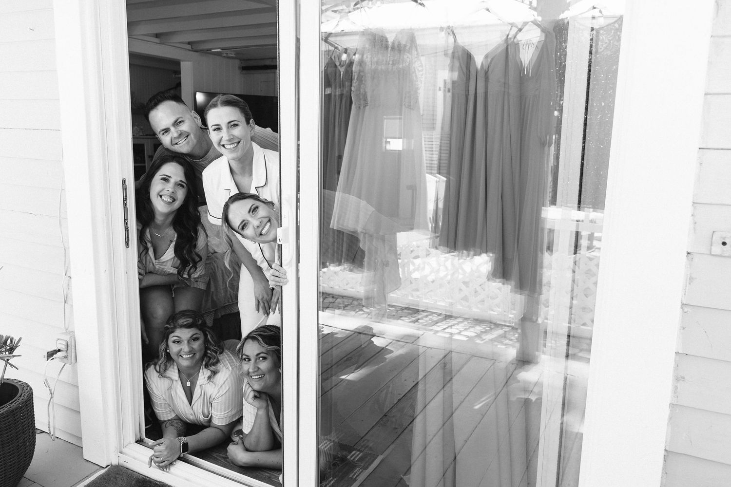 Black and white photo of bridal party peeking through a doorway with bridesmaid dresses reflected in glass.