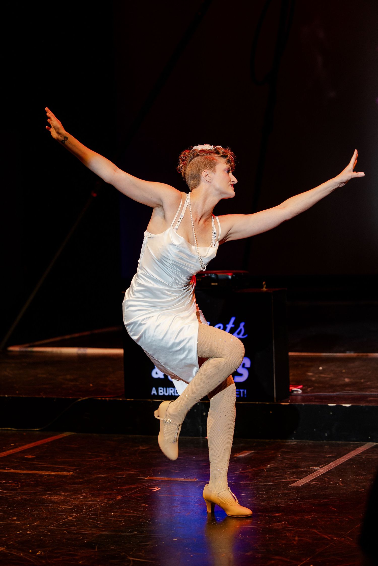 A dancer in a white dress performs on stage with arms outstretched and one leg raised during a burlesque cabaret show.