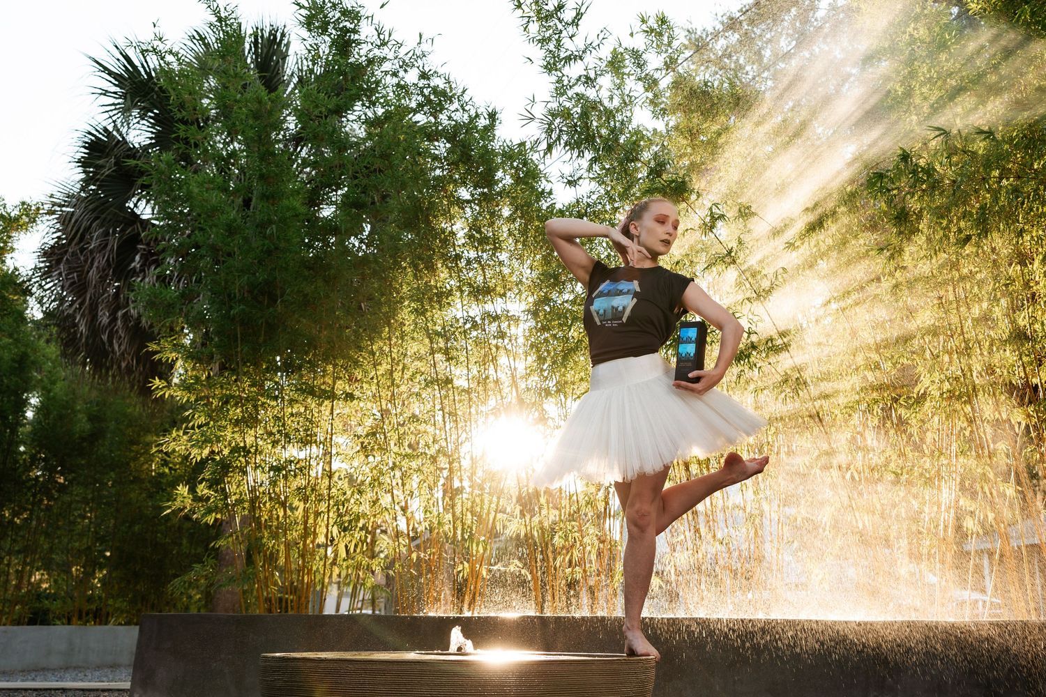 Barefoot dancer in white tutu and black top poses elegantly outdoors with sunlight streaming through bamboo.