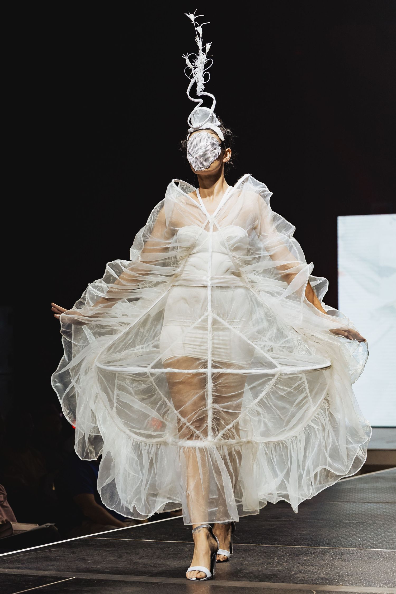Model in voluminous white tulle gown with structured cage skirt and floral headpiece walks the runway.