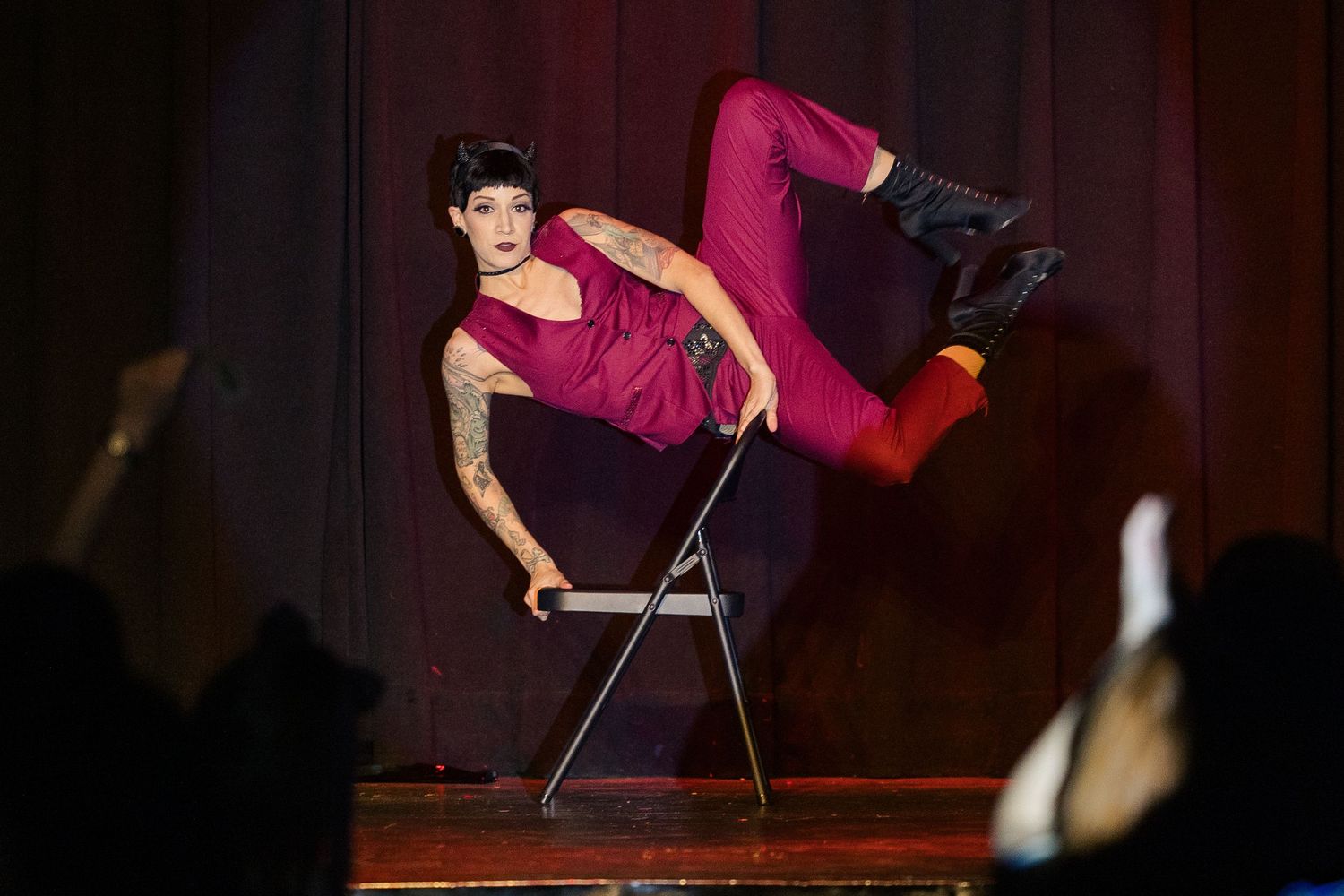 A tattooed performer in a red jumpsuit performs an acrobatic chair act on stage.