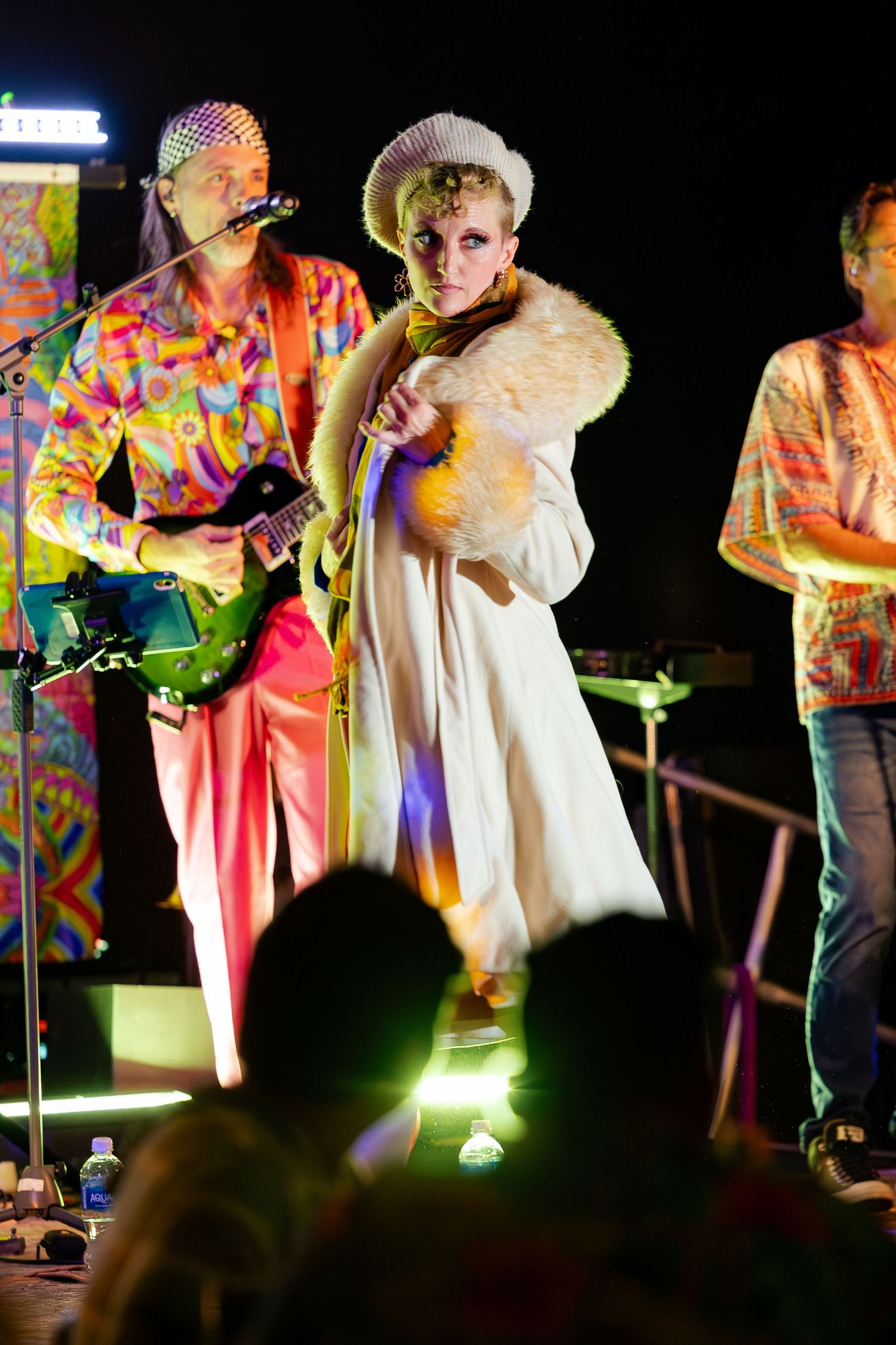 Blonde performer in white fur coat on stage with guitarist in colorful psychedelic outfit behind her.