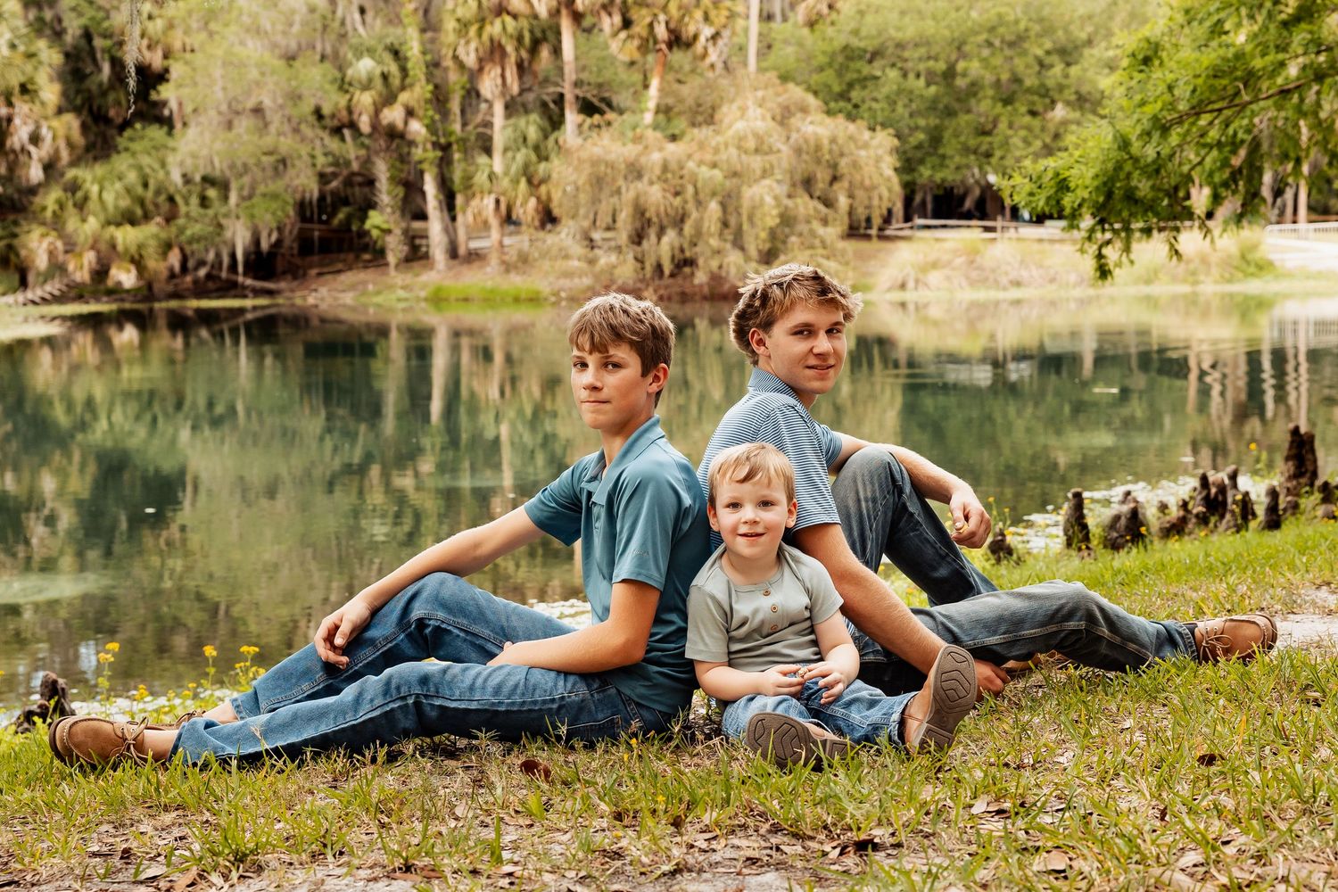 Three brothers sitting together by a serene pond, two older boys and a toddler in casual blue outfits.