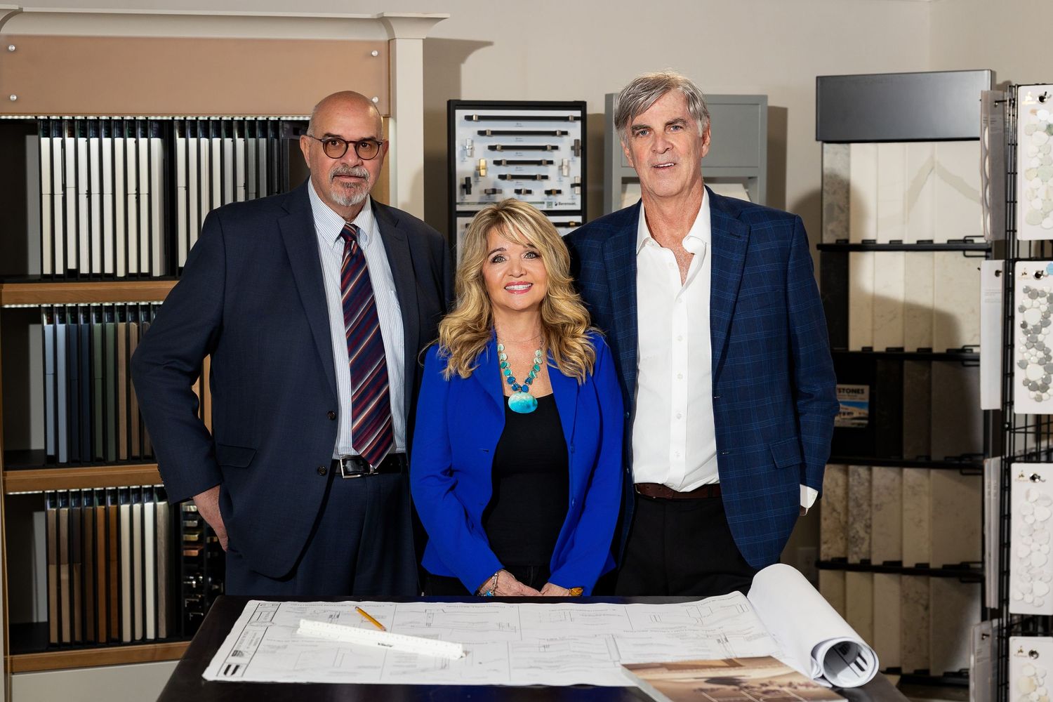 Three professionals pose in a tile and flooring showroom with blueprints and material samples on display.