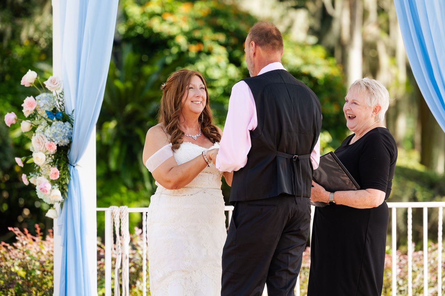 Bride and groom exchange vows at outdoor wedding ceremony with blue draping and floral decorations.