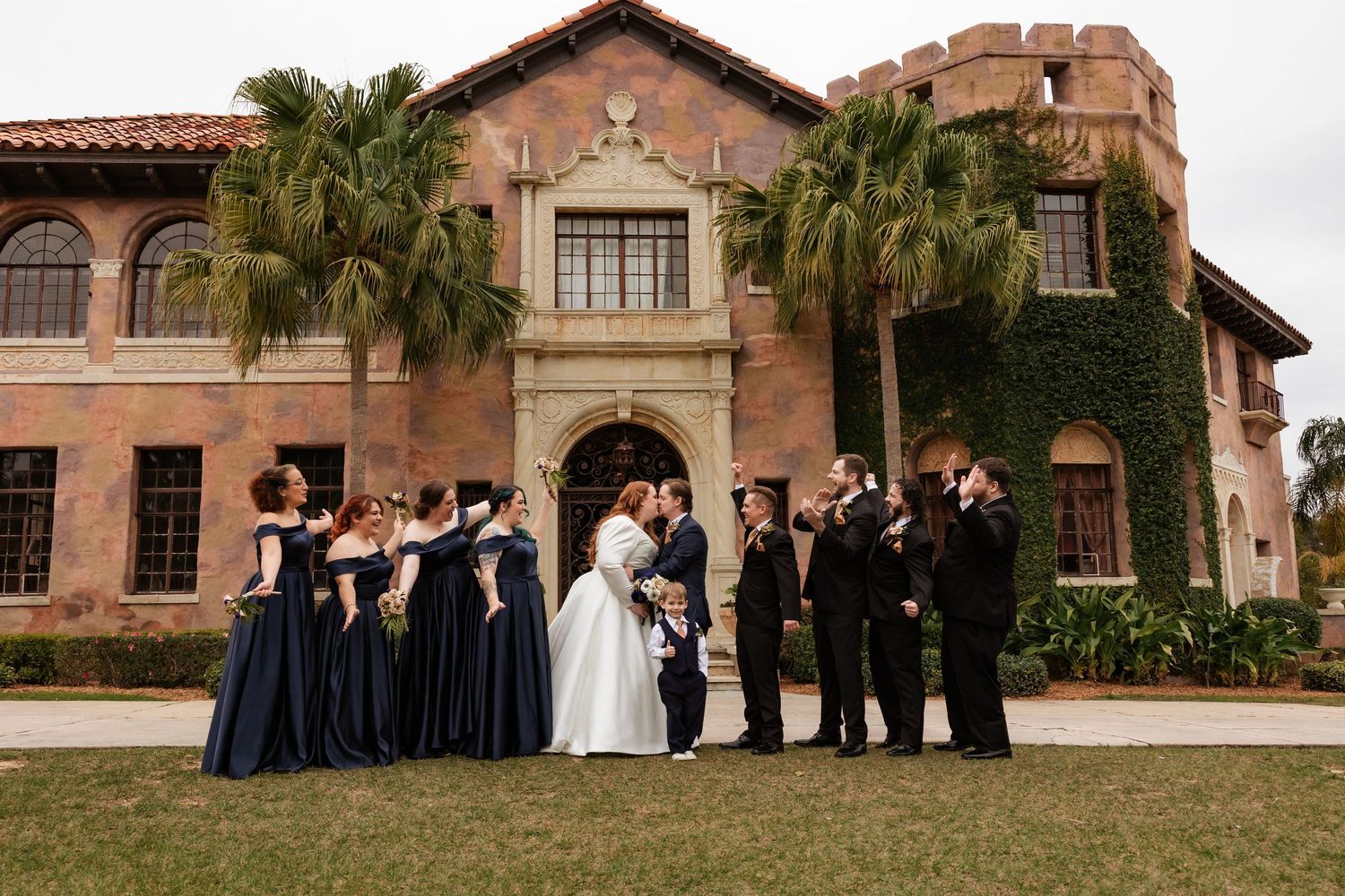Wedding party in navy and black poses kissing in front of Spanish Mediterranean estate with palms.