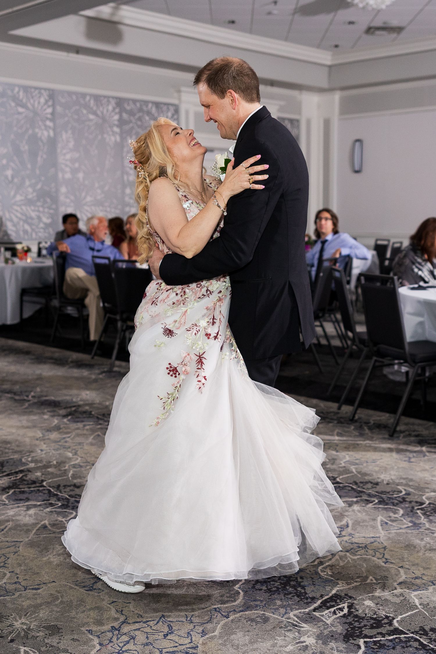 Bride and groom share their first dance in a decorated ballroom with wedding guests seated nearby.