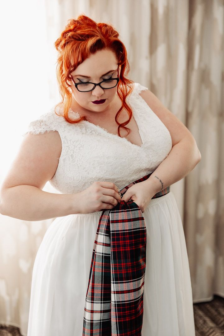 Redhead bride in white lace dress adjusting a plaid tartan sash around her waist before wedding.