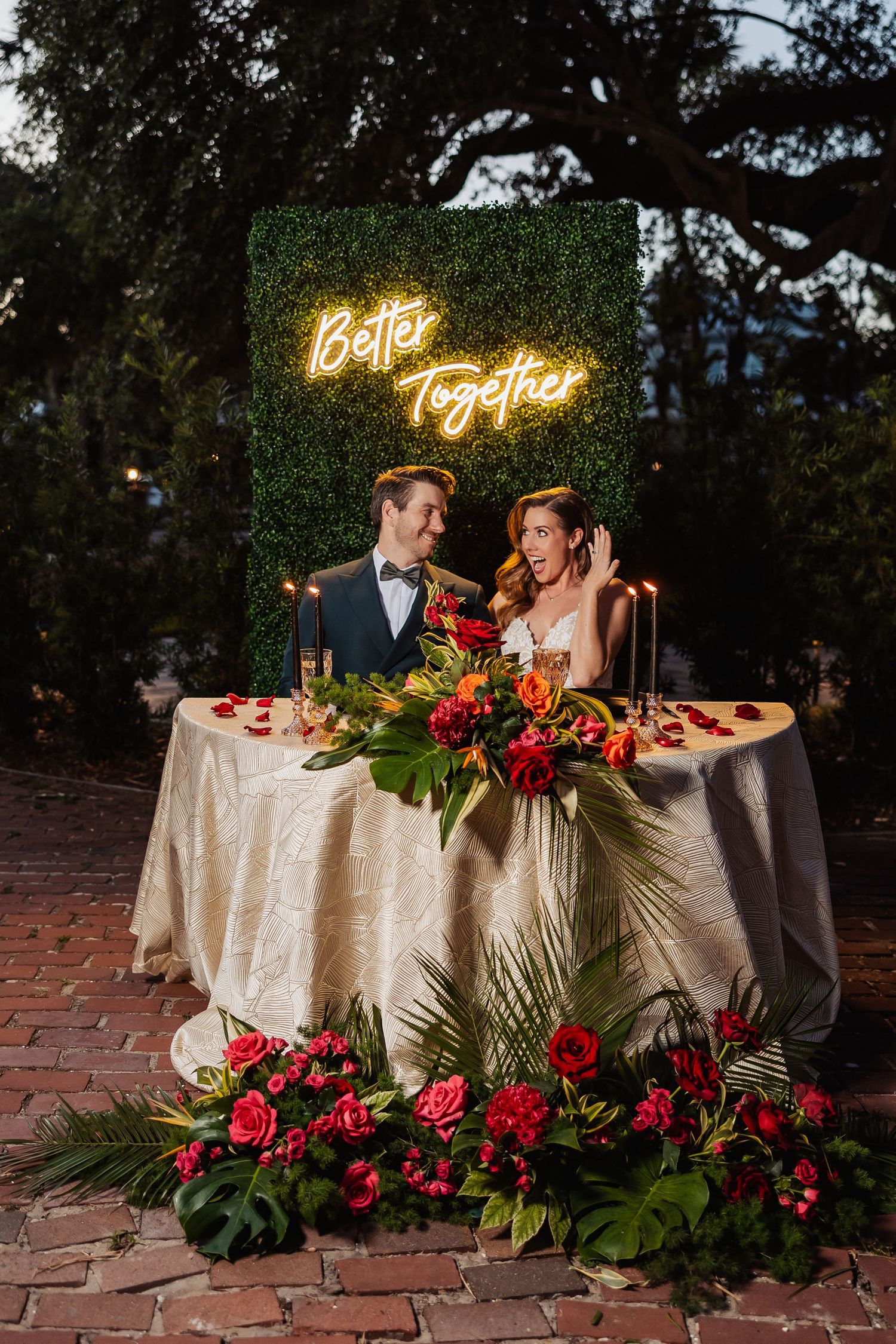 Newlyweds at sweetheart table under Better Together neon sign outdoors.