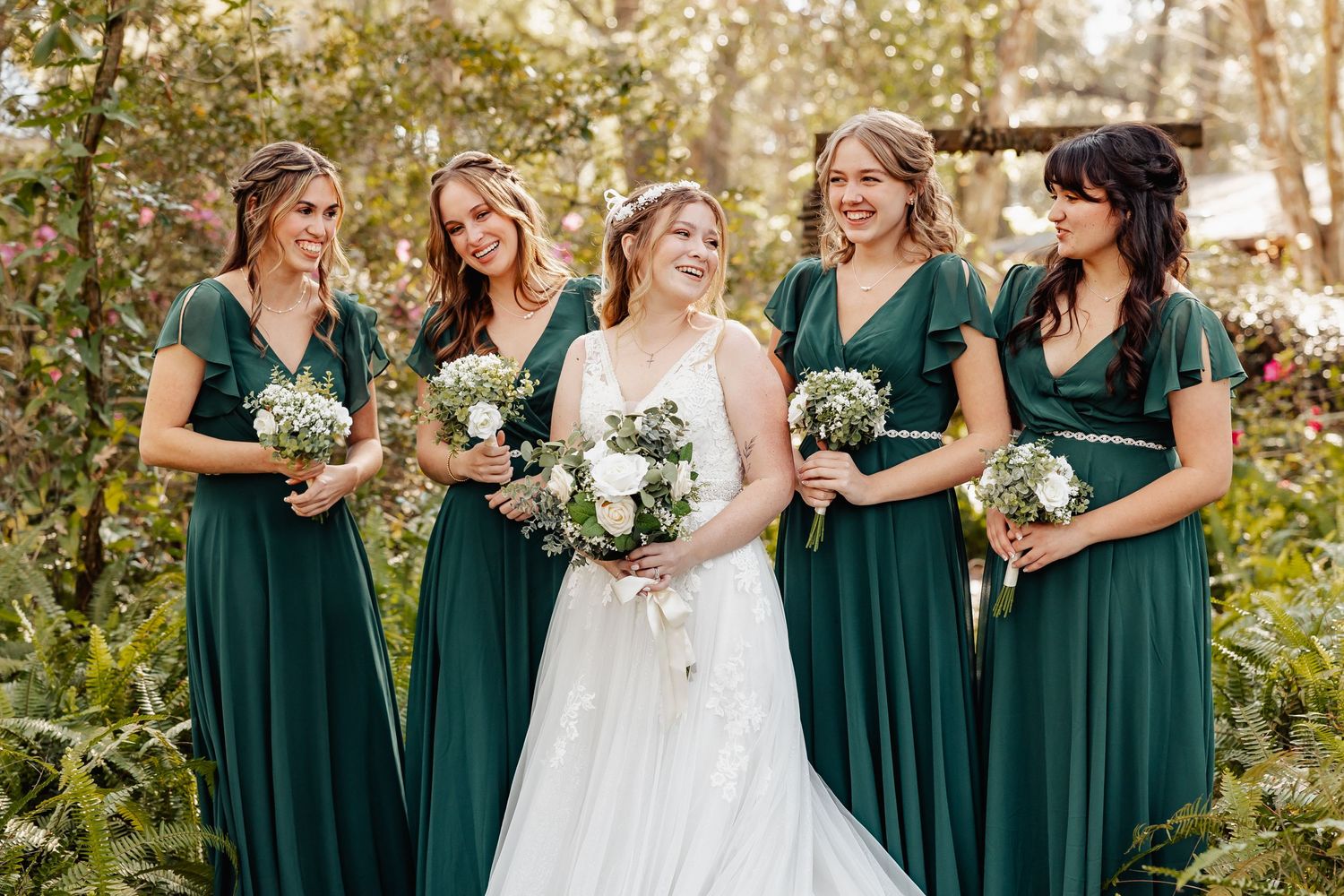 Wedding party in emerald green bridesmaid dresses posing outdoors with floral bouquets.