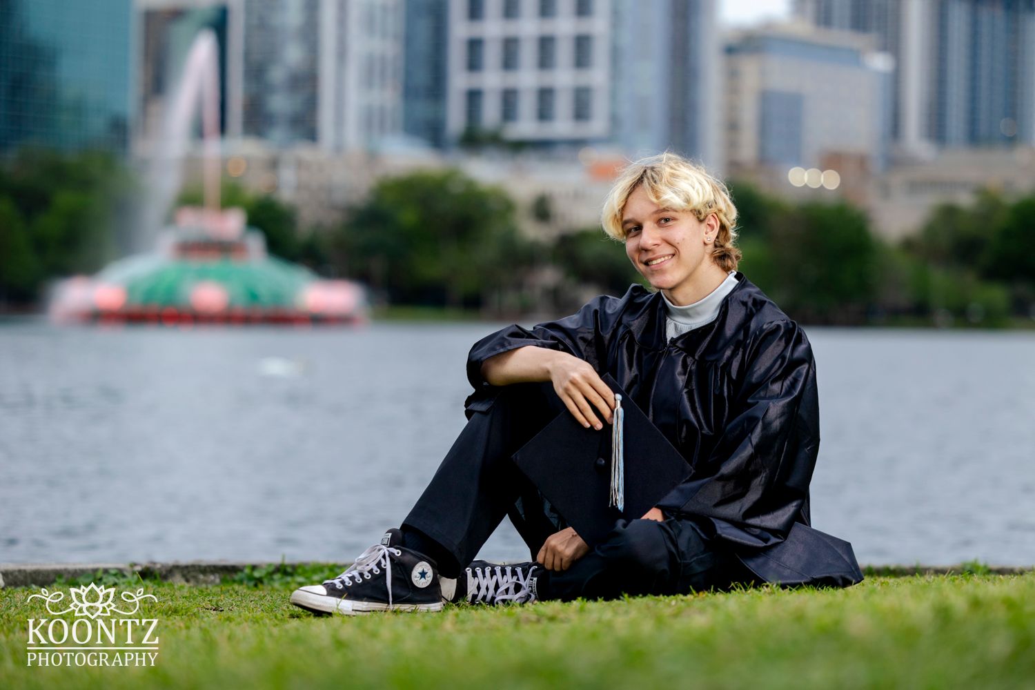 Casual graduation photo on grass with city skyline and fountain in background.