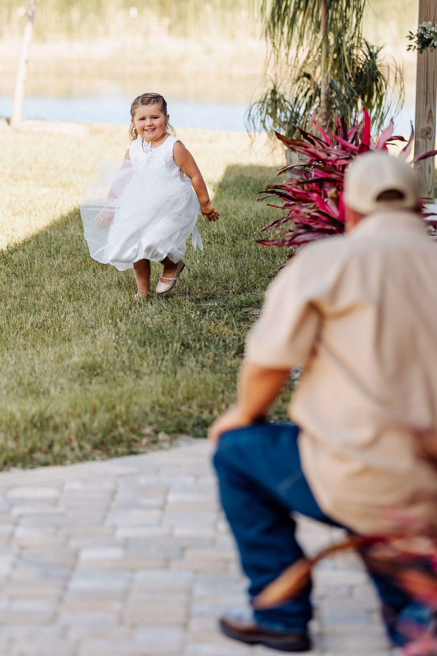 A small child in a white dress runs playfully on grass near tropical plants while someone watches from a paved area.