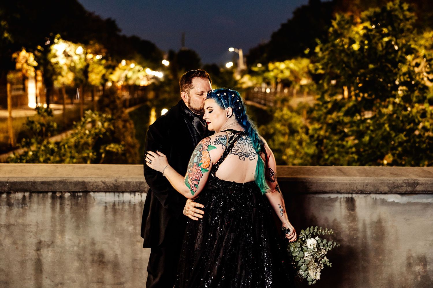 Gothic bride in black gown with blue hair and tattoos embraced by groom at night outdoor wedding.