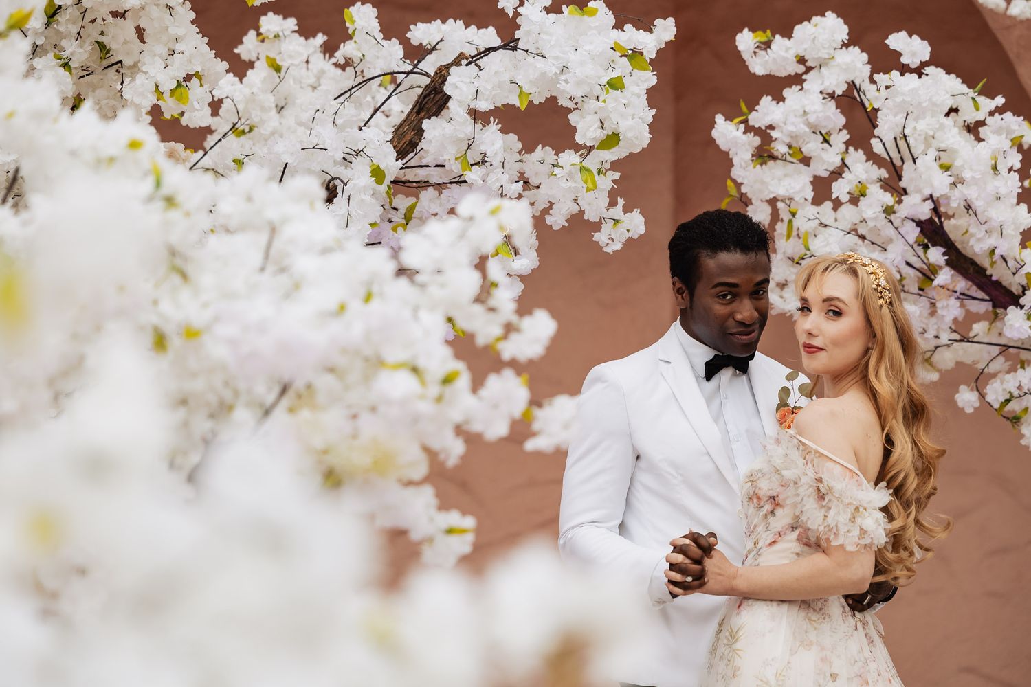 Elegant couple in wedding attire embracing beneath blooming white cherry blossoms against terra cotta wall.