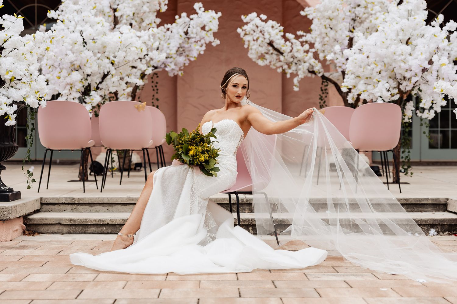 Elegant bride in white gown poses on terrace with flowing veil and floral decor under cherry blossom branches.