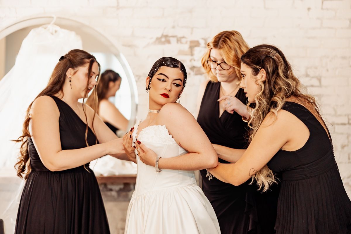 Bridesmaids in black dresses helping bride with strapless white wedding gown in bright white room.