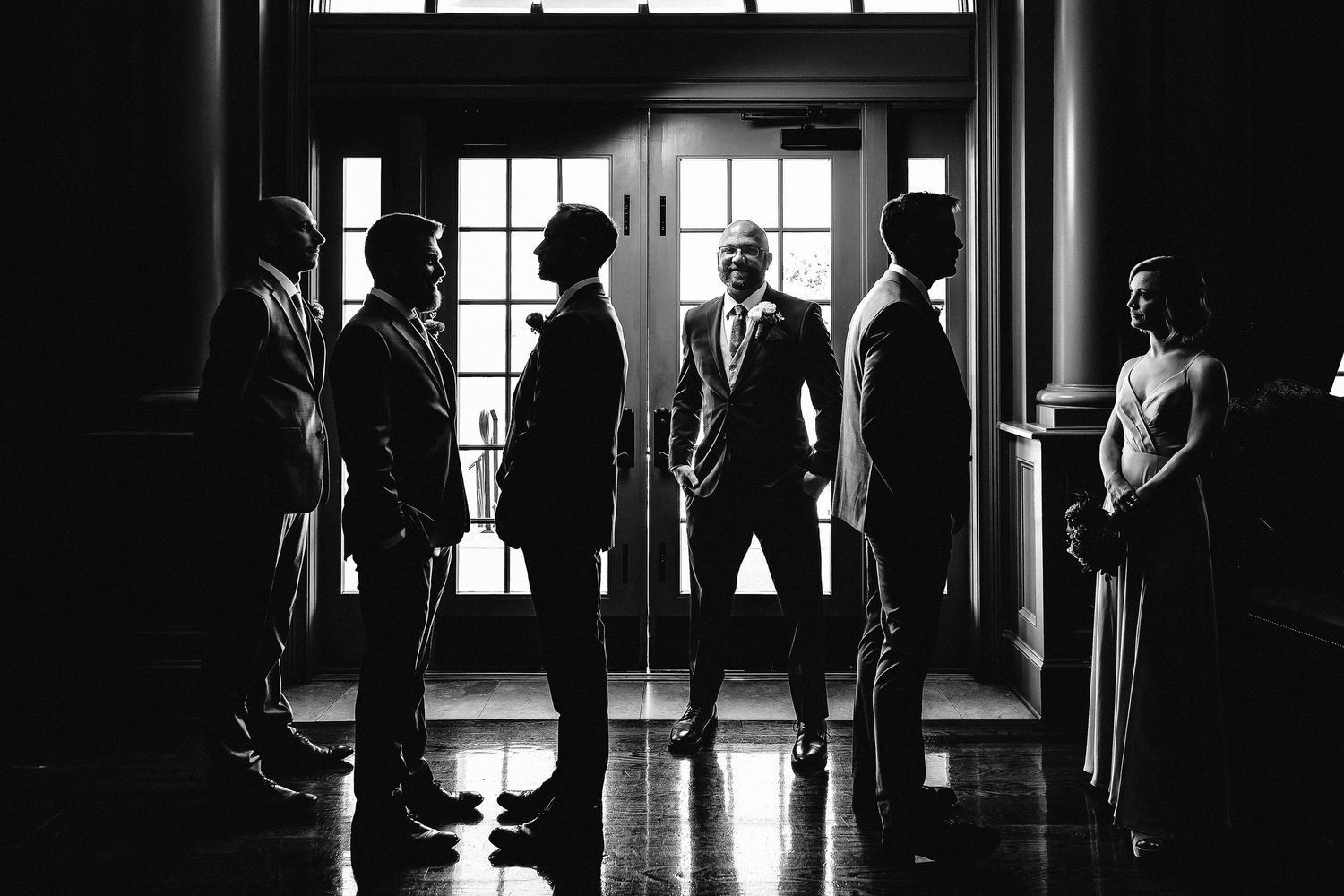 Dramatic black and white silhouette of groom and groomsmen standing in a sunlit doorway.