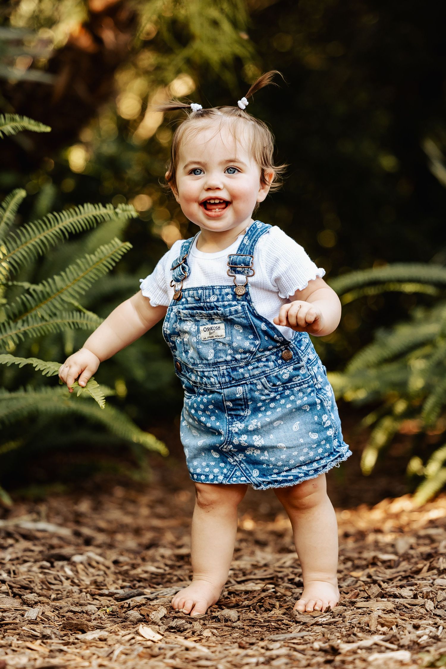 Happy toddler girl in denim overalls stands barefoot smiling in a sunlit garden with ferns.