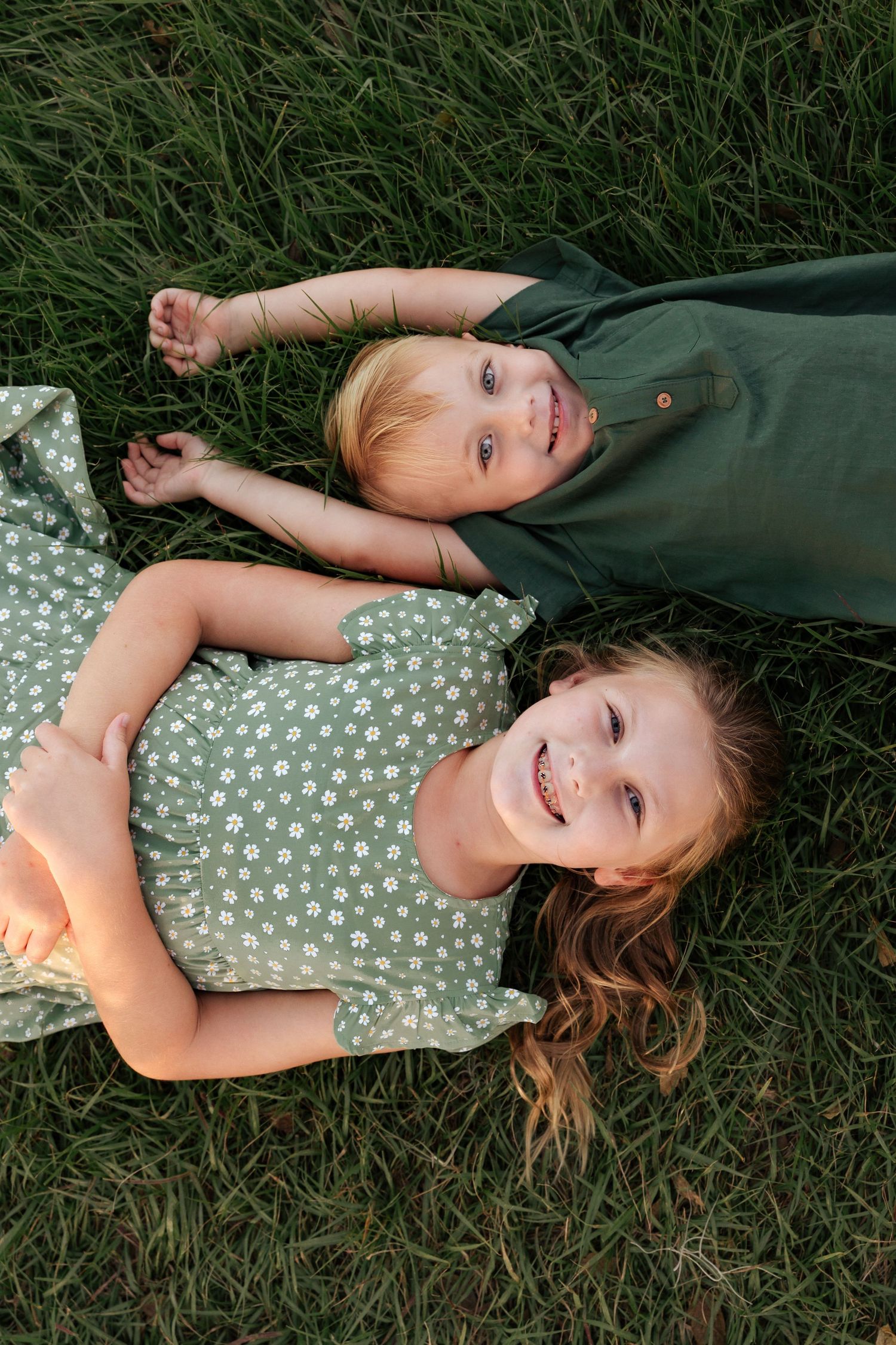 Two smiling children lying on green grass, girl in floral dress and boy in dark green shirt.