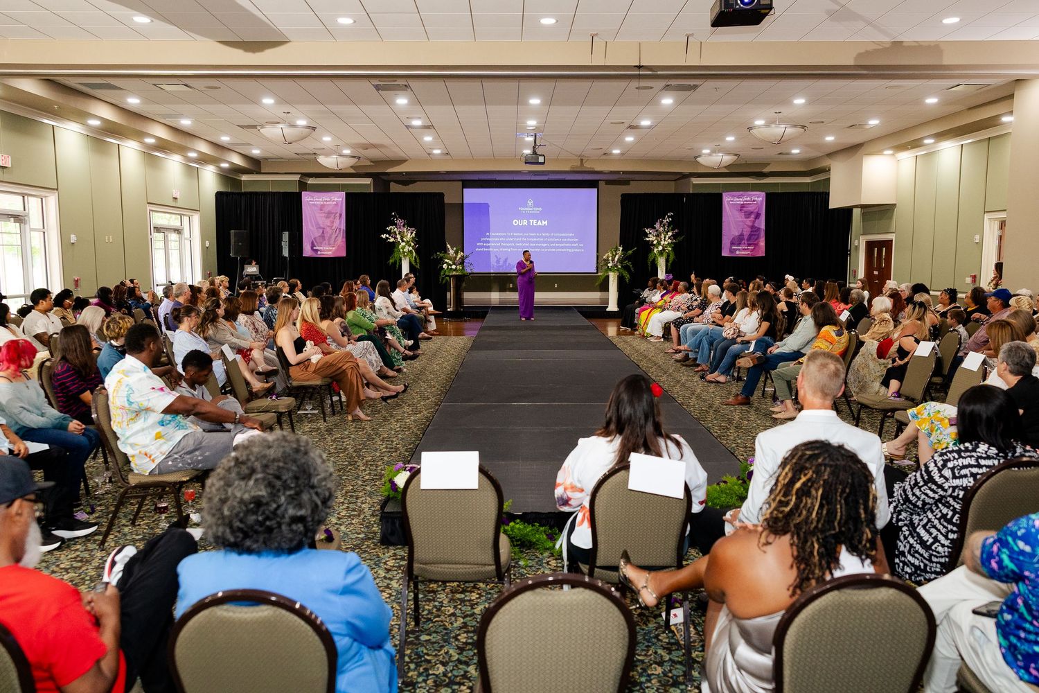 Large audience seated in rows watches a fashion show presentation in an elegant ballroom venue.