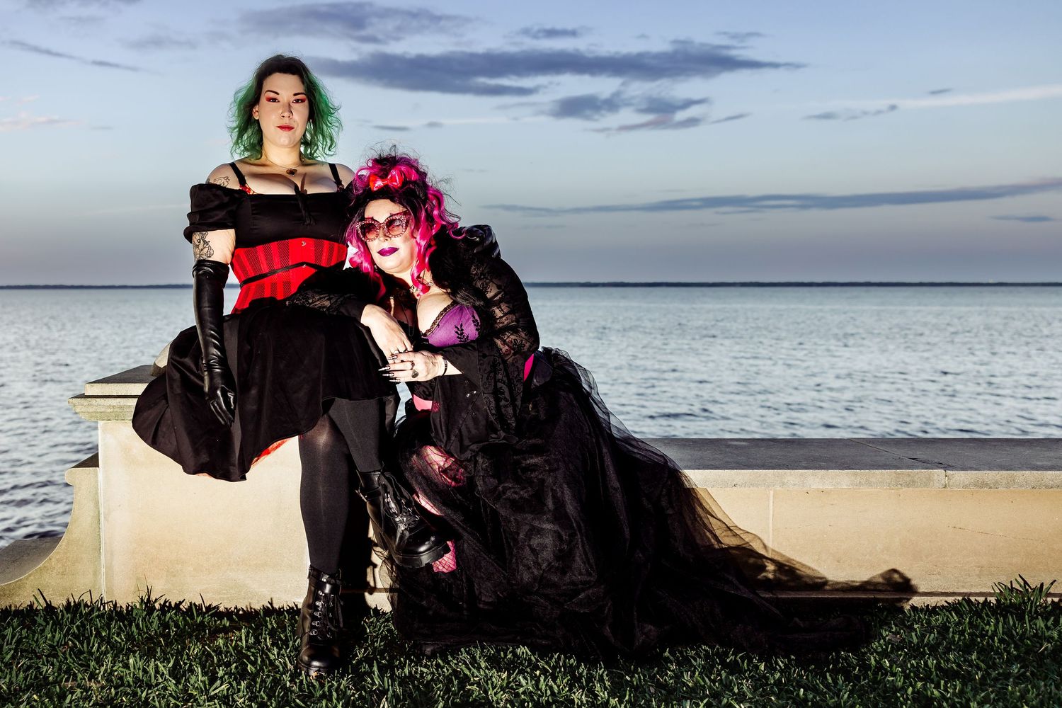 Two women in elaborate gothic black dresses with colorful hair pose outdoors near waterfront at dusk.