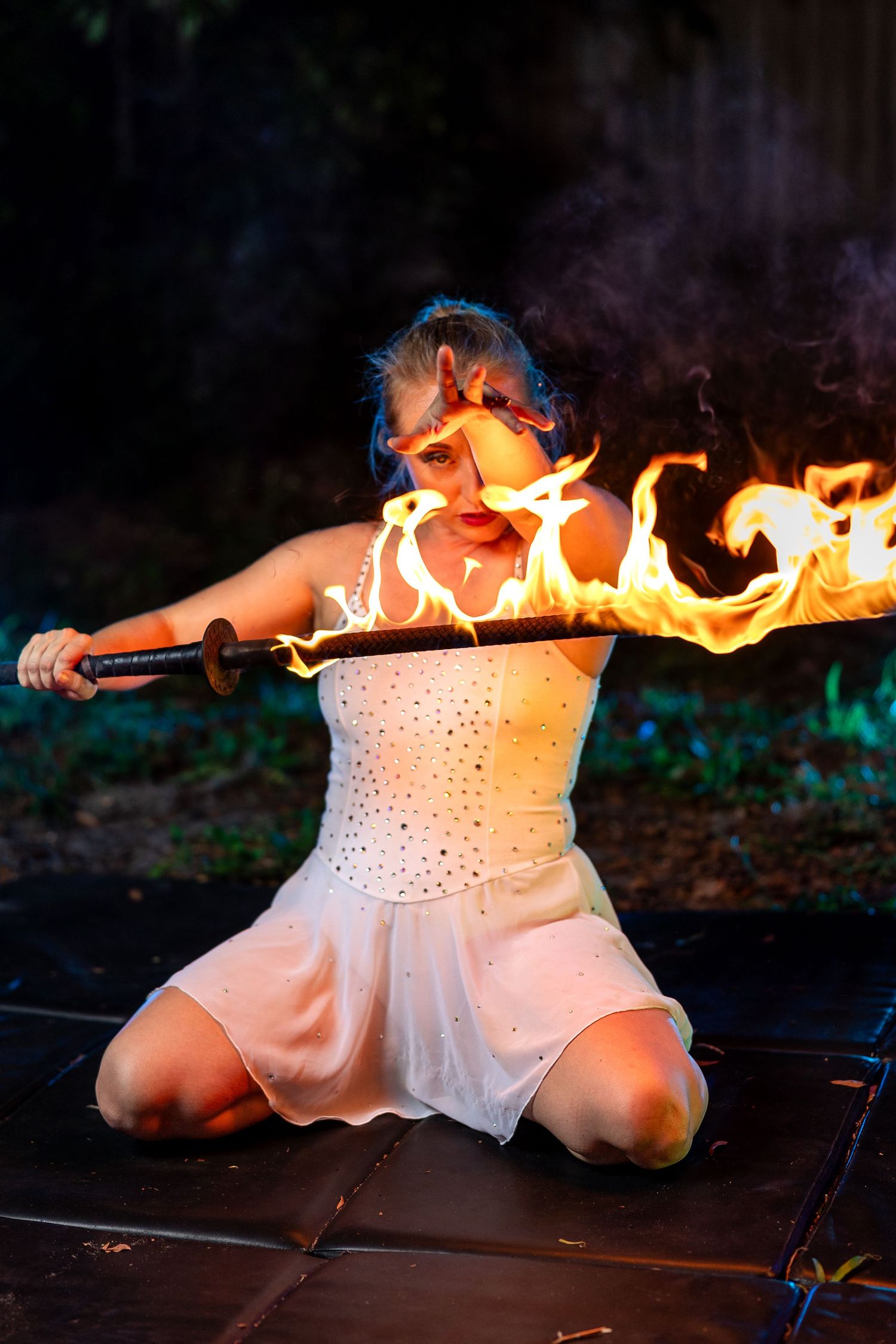 Female performer kneeling and holding a horizontal flaming staff close to her face at night outdoors.