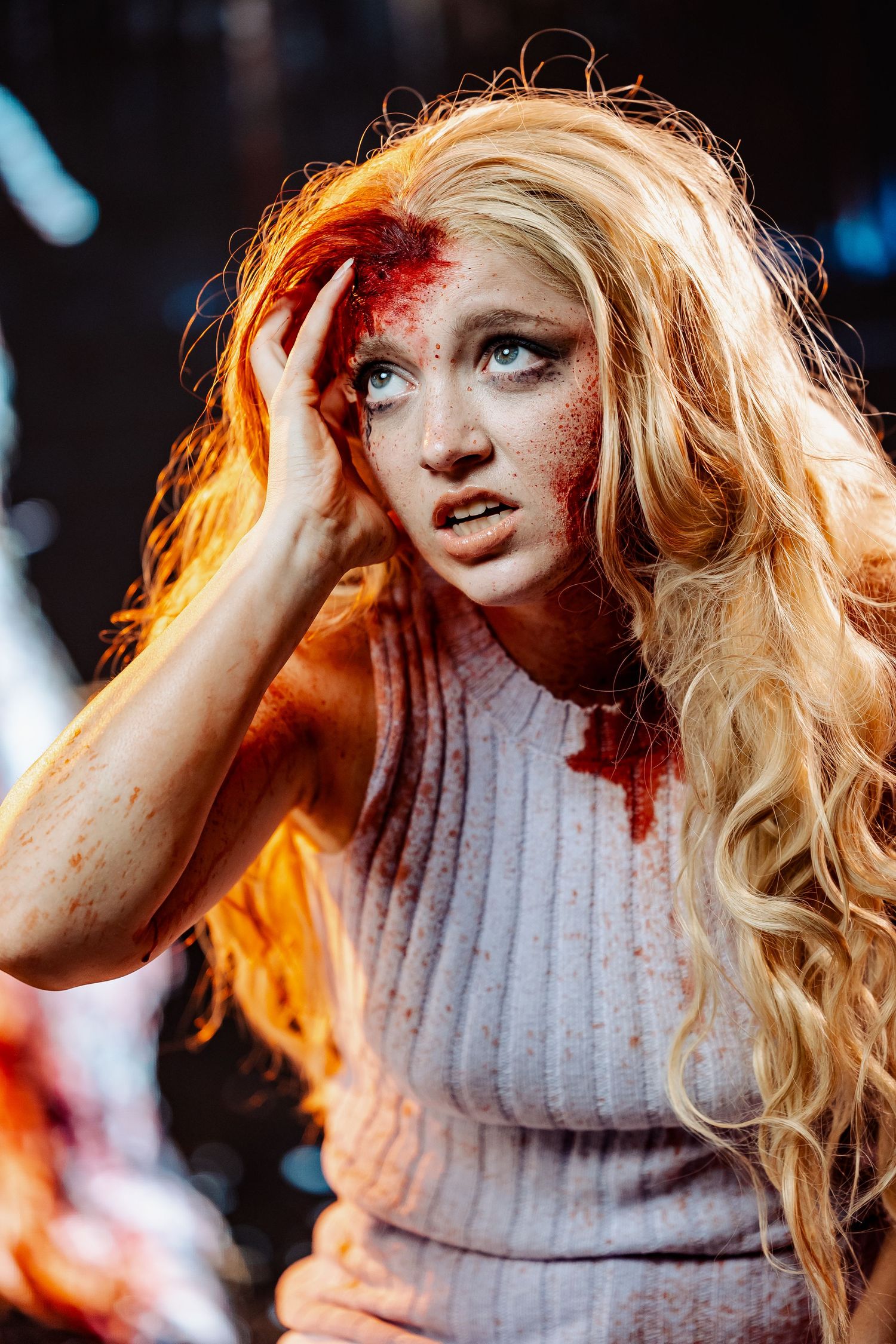 Blonde woman with realistic bloody wound special effects makeup touching her head in dramatic scene.