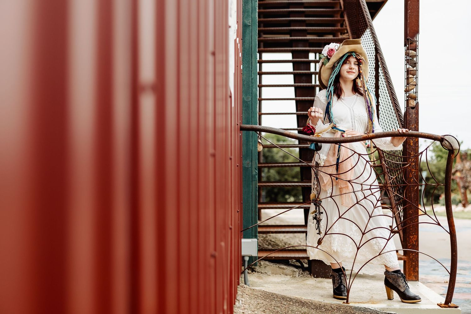 Woman in spiderweb skirt and floral hat stands on red metal staircase railing outdoors.
