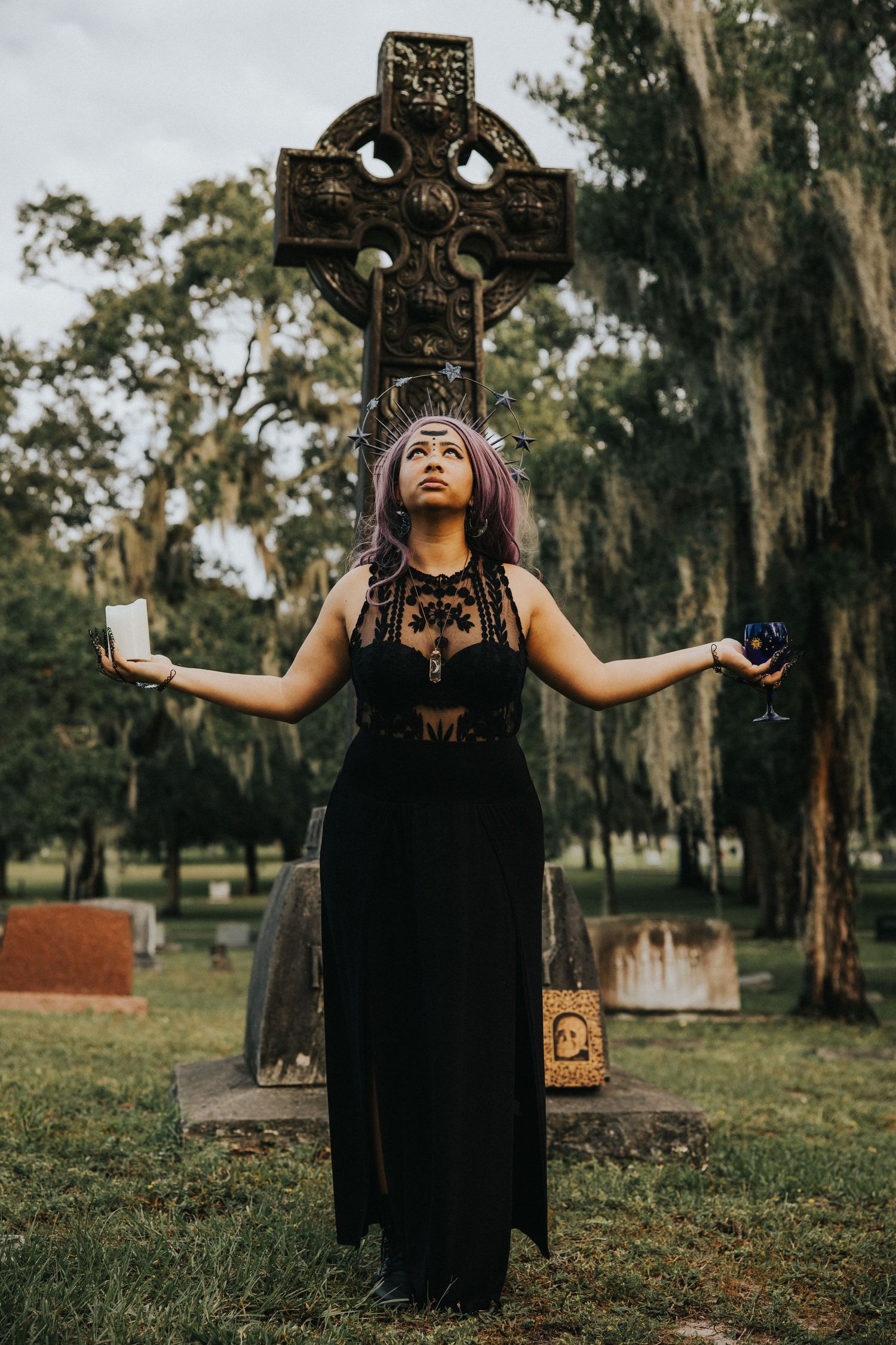 A woman in a black dress stands arms outstretched before a Celtic cross gravestone in a cemetery.