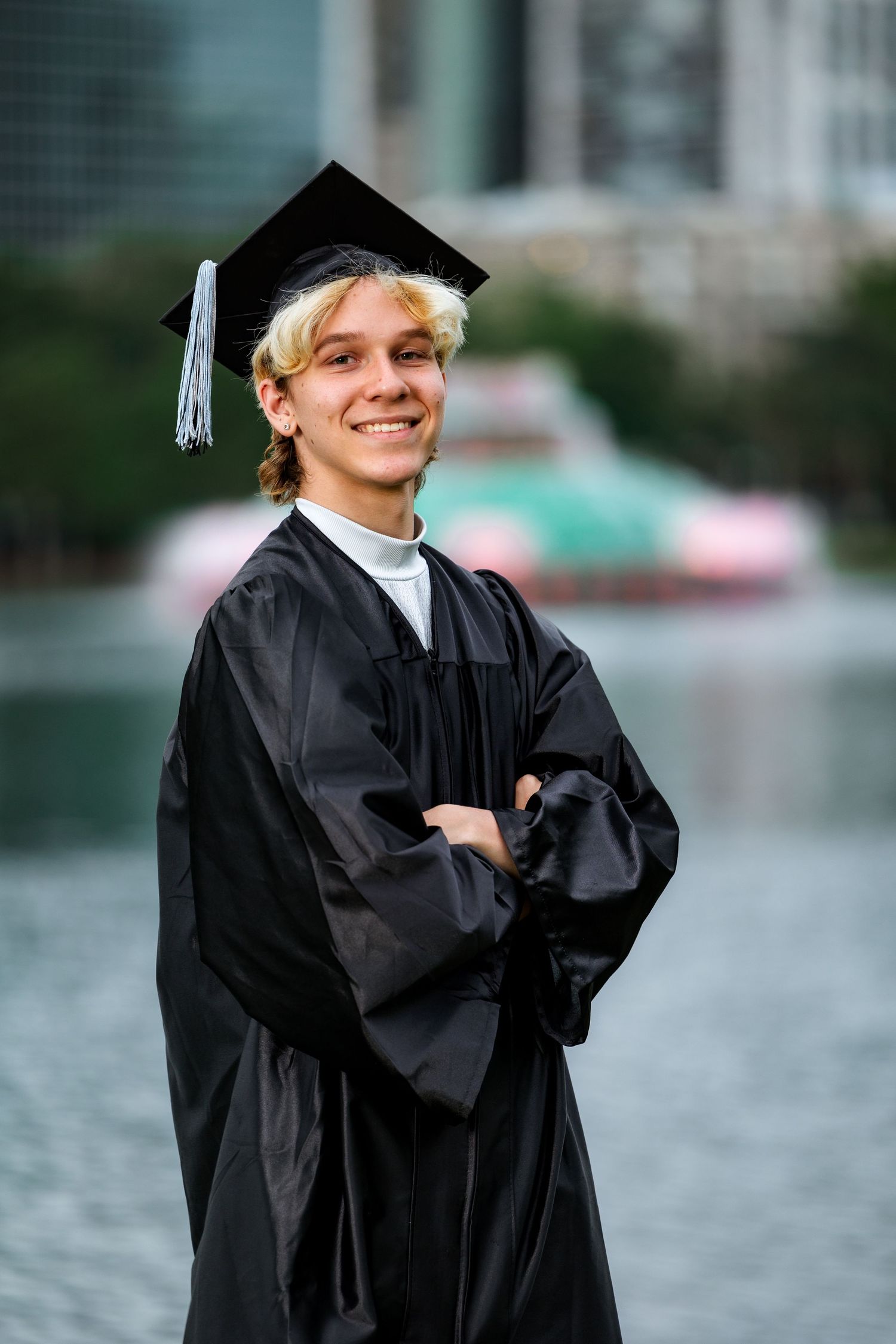 Smiling young graduate in black cap and gown with arms crossed, posing outdoors near a waterfront.