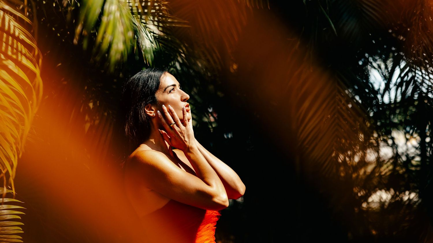 A woman in a red dress poses thoughtfully among lush tropical palm leaves in warm golden sunlight.