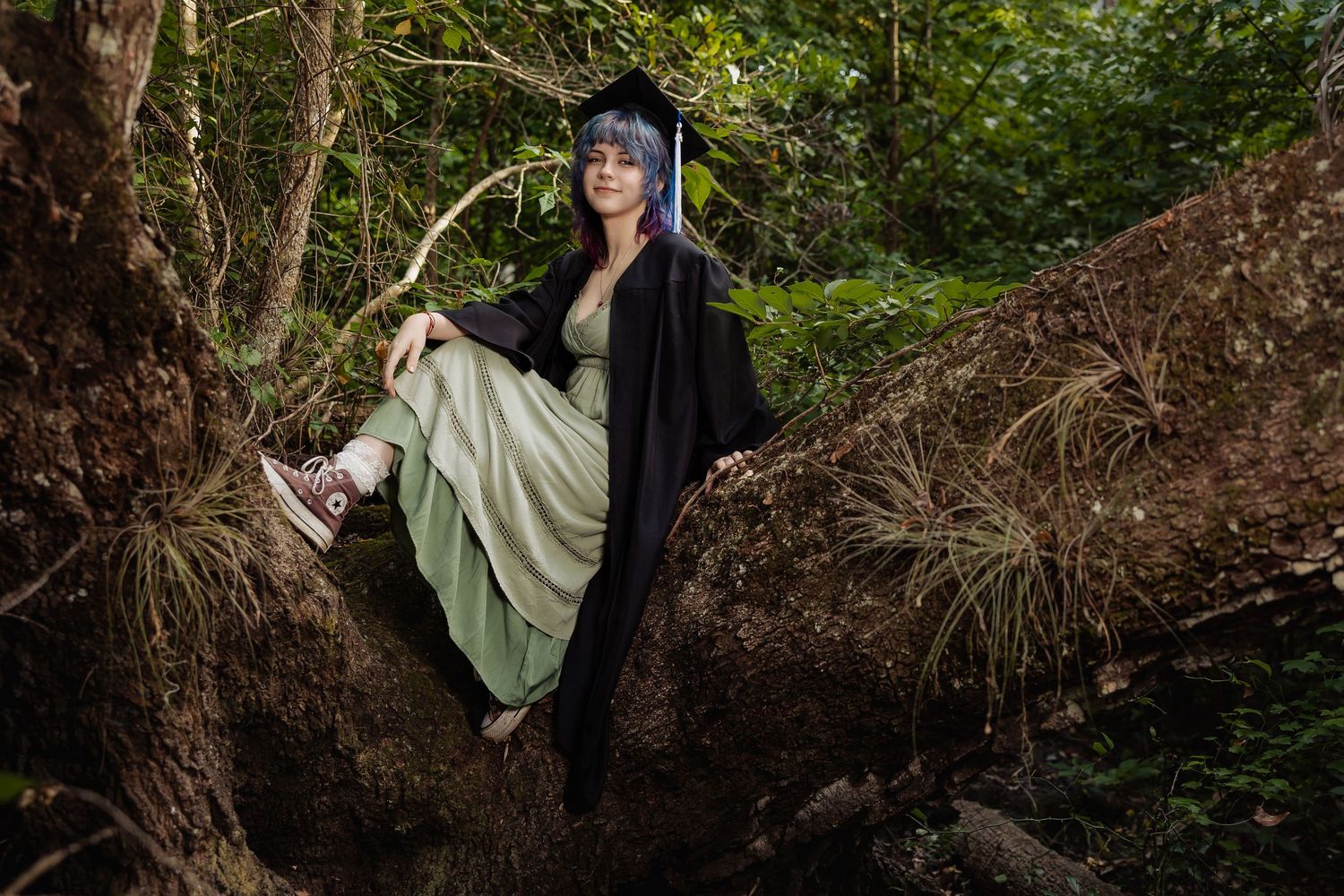 A woman in graduation cap and gown sits on tree roots in a lush green forest wearing a sage dress and sneakers.