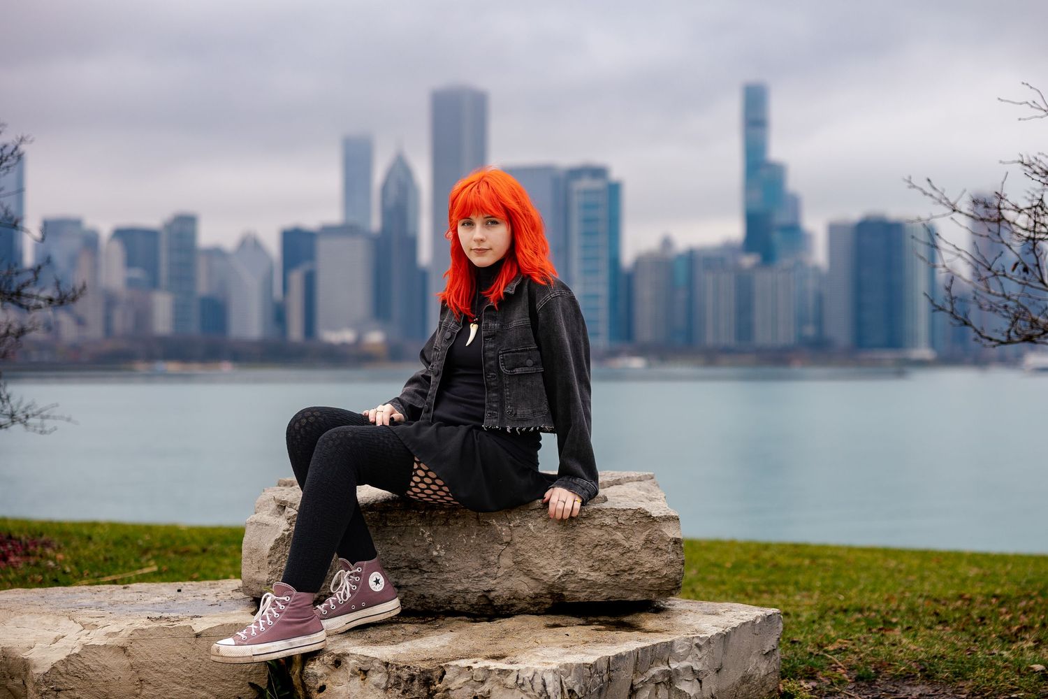 Young woman with vibrant orange hair sits on rocks with Chicago skyline and lake in background.