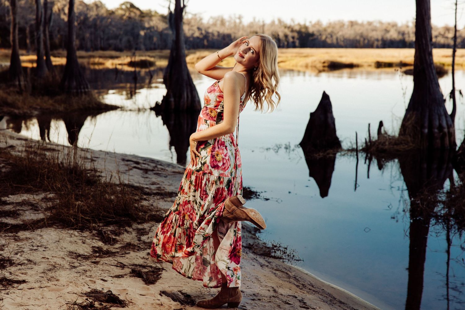 Woman in floral maxi dress and boots posing by a serene swamp with cypress trees in golden light.