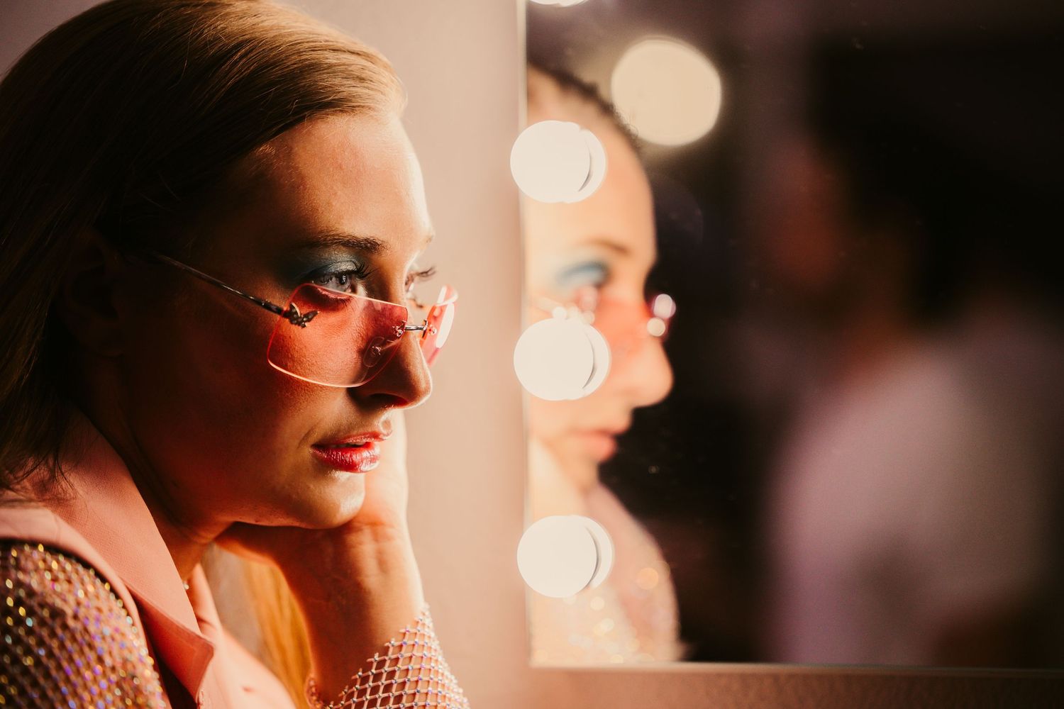 Blonde woman with pink-tinted glasses gazes into a Hollywood vanity mirror with warm bokeh lights.