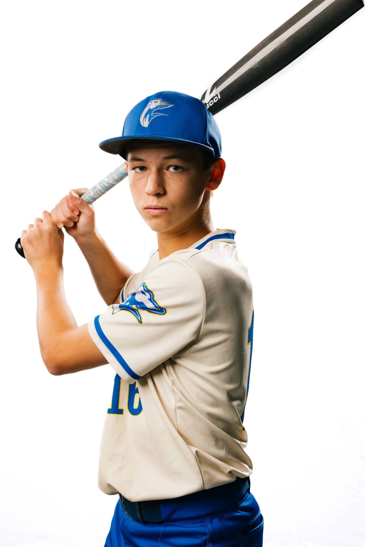Young baseball player in blue helmet and cream uniform poses with bat in batting stance against white background.