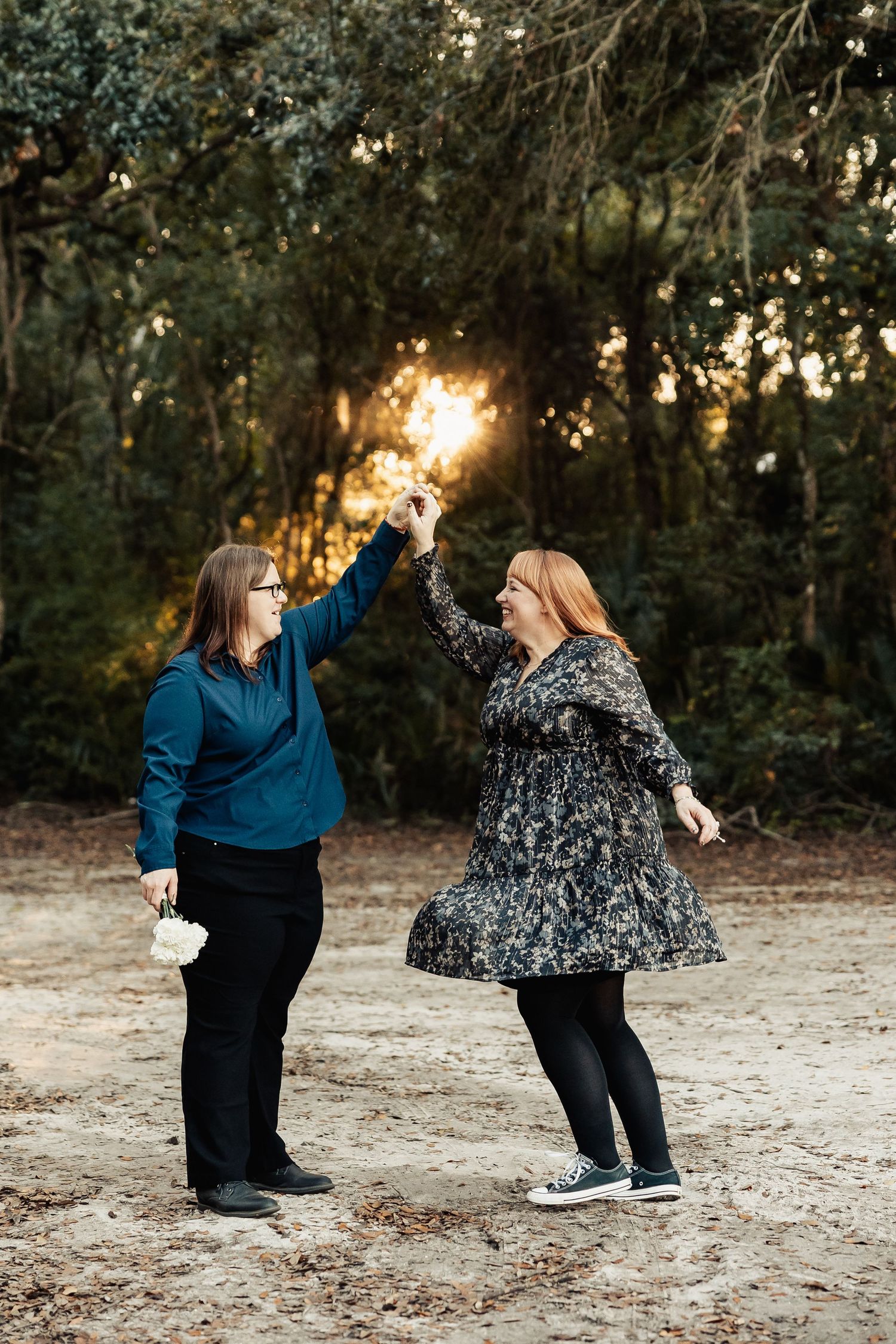 Two women dance joyfully outdoors at sunset, holding hands raised in a wooded park during an engagement session.