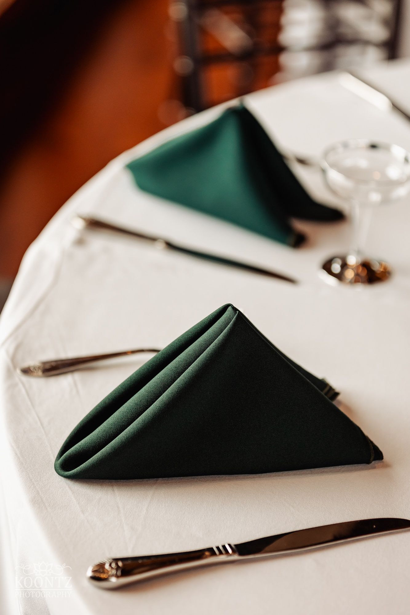 Elegant dark green napkin folded on white tablecloth with silverware at formal place setting.