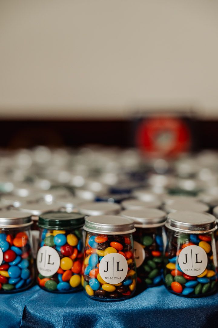 Rows of small glass jars with JL monogram labels filled with colorful M&Ms candies on blue fabric.
