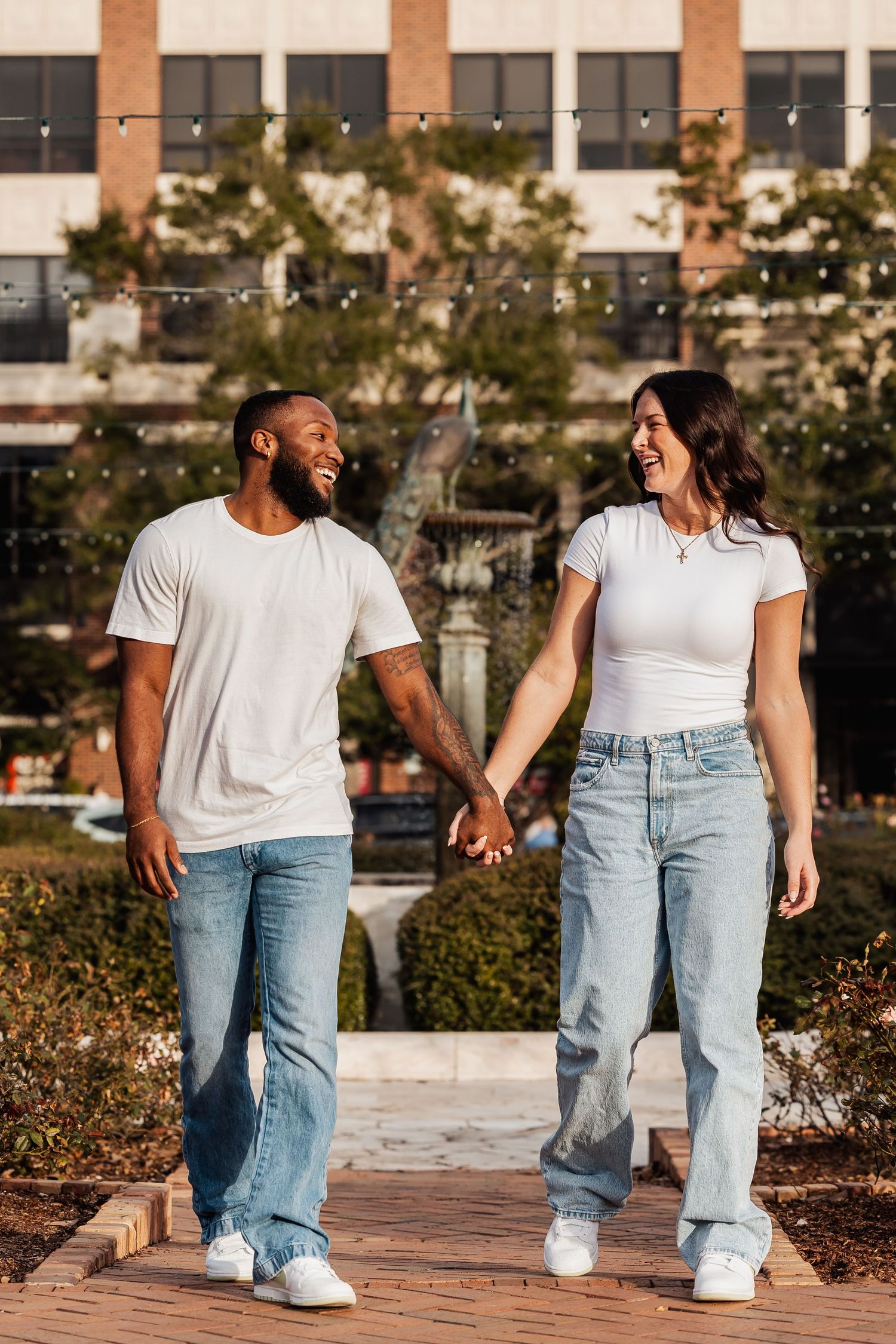 A joyful couple holds hands while walking together on a brick pathway in warm sunlight.