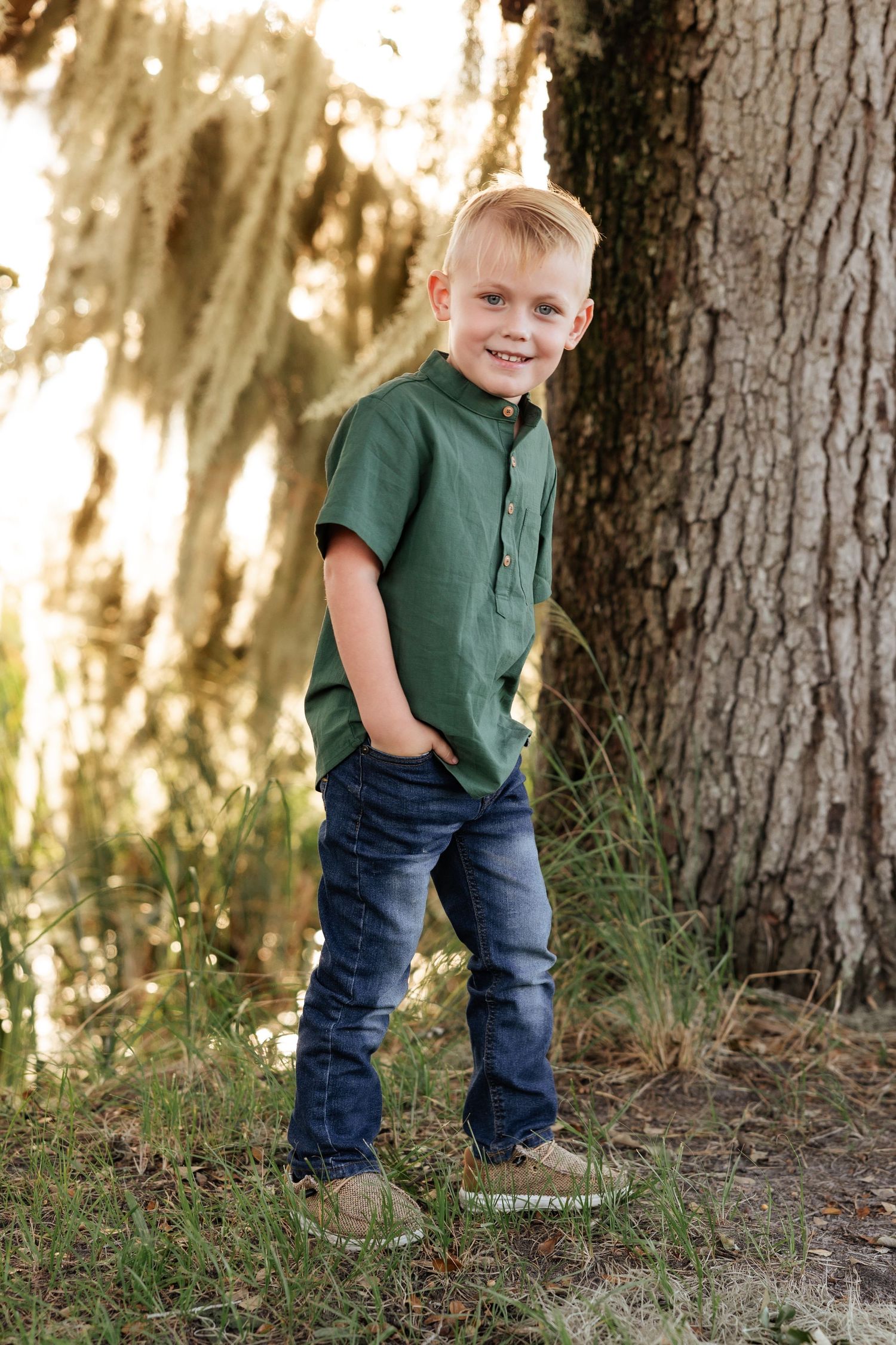 Child poses casually with hands in pockets next to large tree trunk.