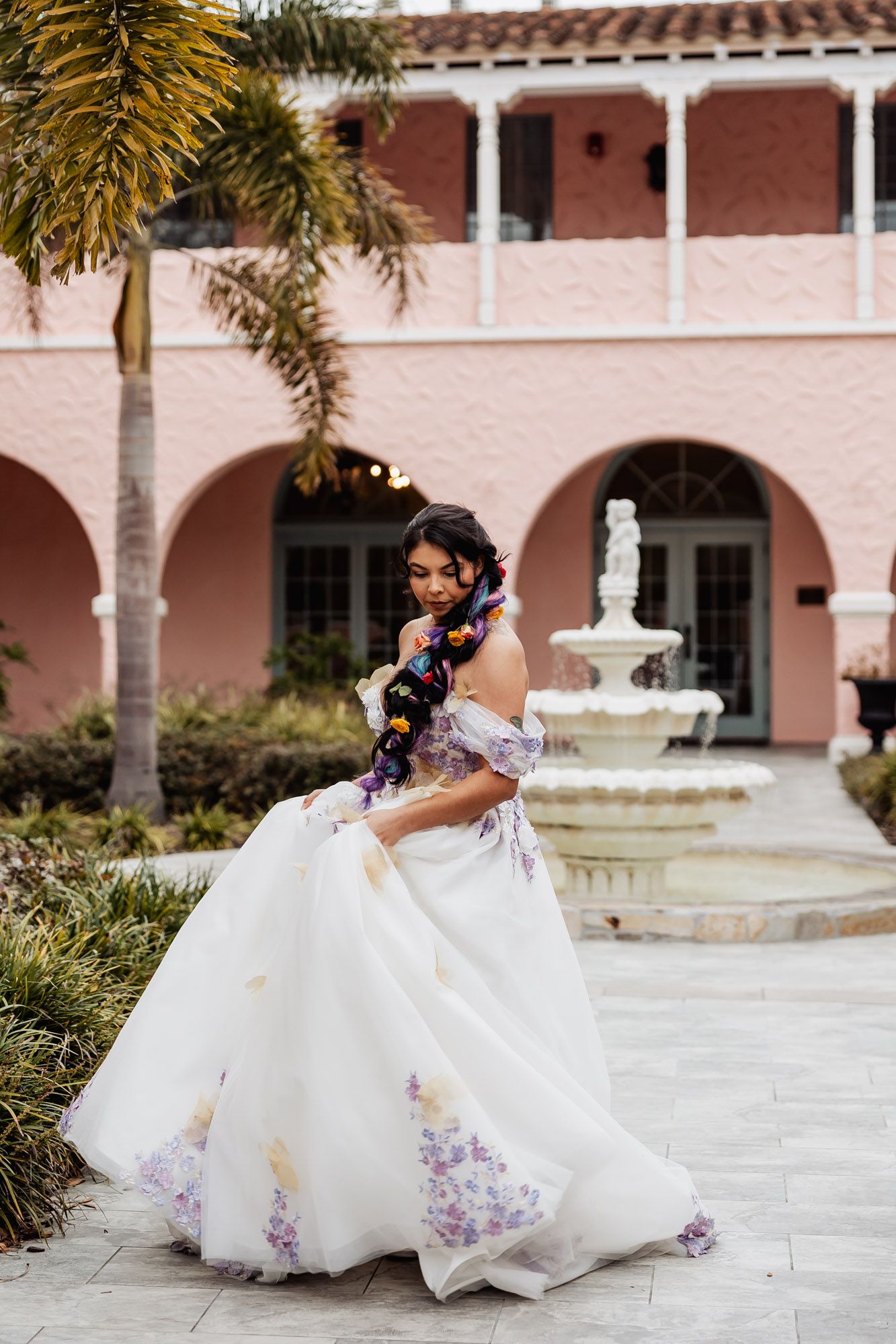Bride twirls in white floral ball gown with colorful hair accessories near fountain at pink estate.