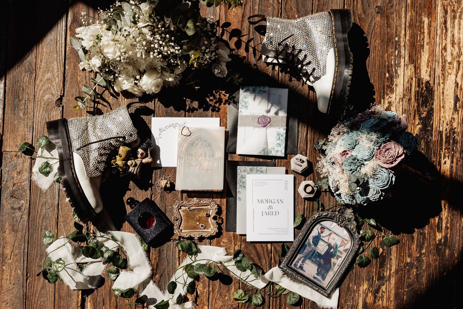 Elegant gothic wedding flat lay with invitations, rings, boots, and floral arrangements on rustic wood.