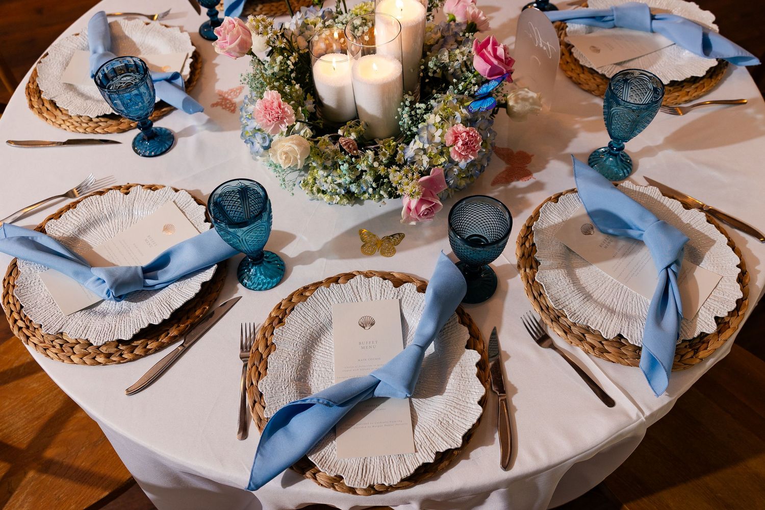 Elegant wedding reception table setting with blue goblets, wicker chargers, and floral centerpiece with white candles.